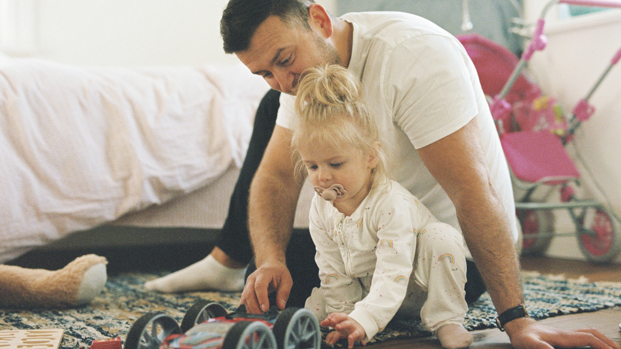 En man och en liten flicka sitter på golvet i ett barnrum och  leker med en bil. 