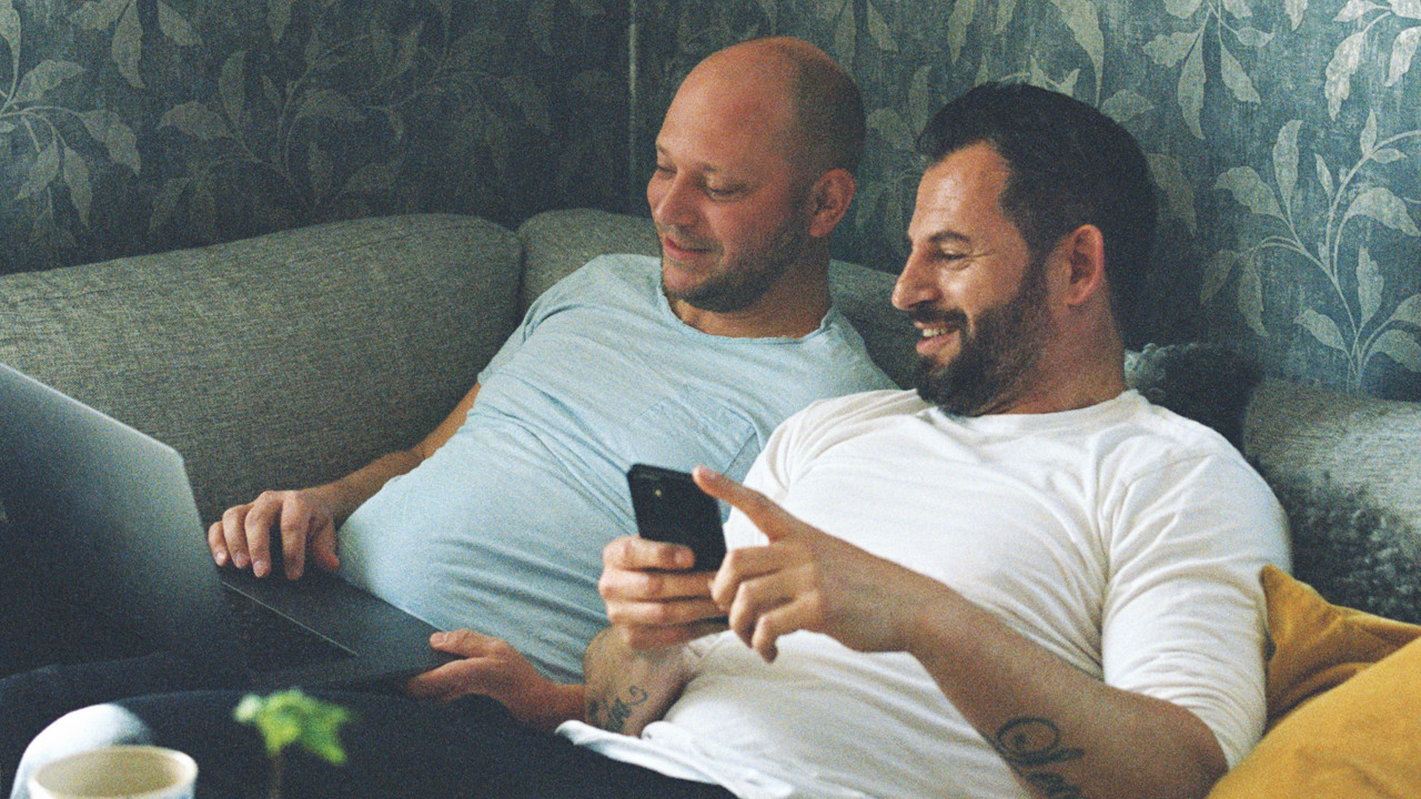 Två män umgås i soffan medan de tittar på laptop och mobil