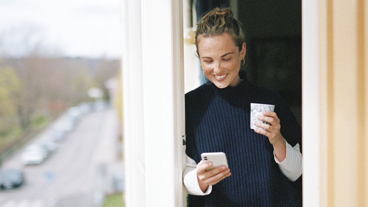 Ung kvinna ler medan hon står på sin balkong med en kaffekopp i handen och tittar på sin mobil