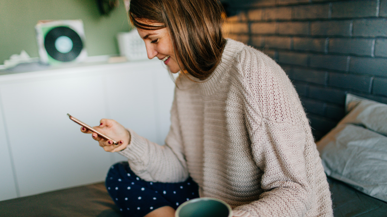 Ung kvinna ler  medan hon sitter i sängen med en kopp kaffe och tittar på sin mobil